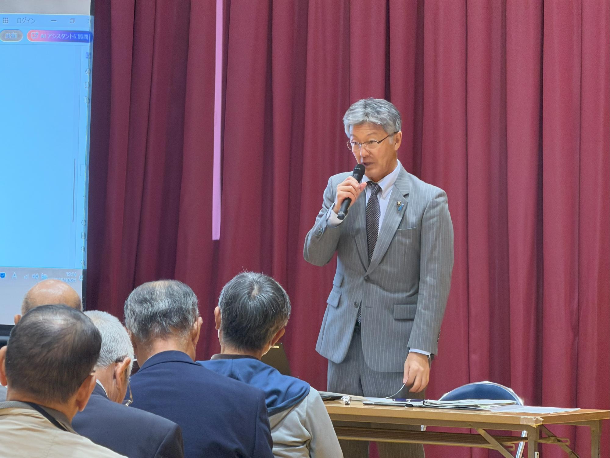 写真：マイクを持ち、立ち上がって住民に挨拶をする市長