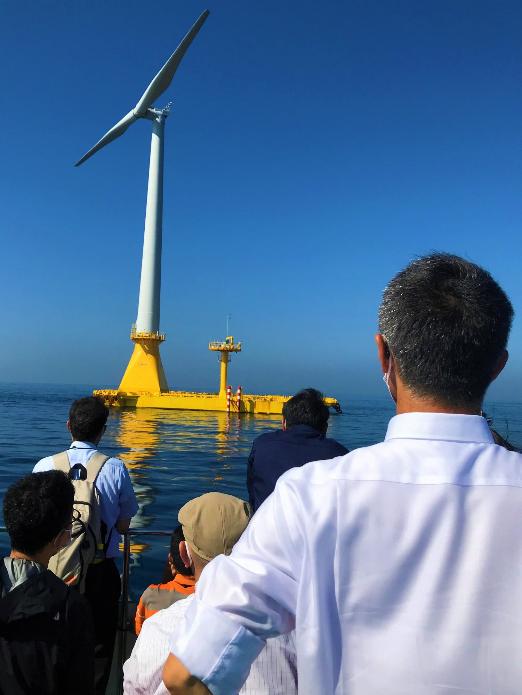 海に浮かぶ黄色と白色の浮体式洋上風力設備を見ている市長の後ろ姿の写真