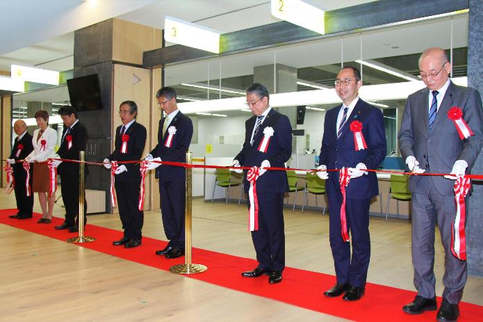 新庁舎1階執務室を背景に、赤いカーペットが敷かれている上で、市長と他来賓7名が一列に並んでテープカットをしようとしている写真
