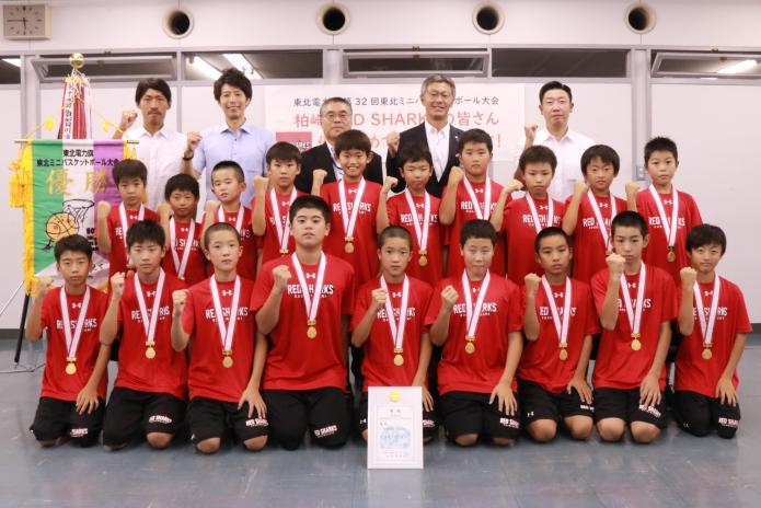 赤いユニフォームを着て、首からメダルをかけた子どもたちがコーチや市長らと集合写真を撮っている様子
