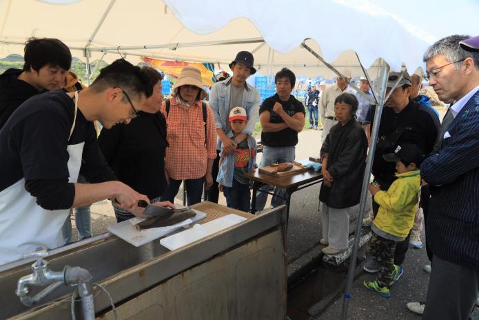 魚を包丁でおろしているところを人々が取り囲んでいる様子の写真