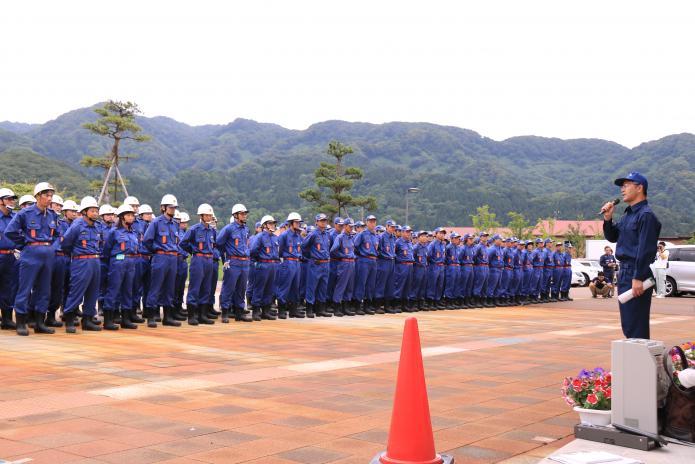 青い制服、白いヘルメットを身に付けて整列する消防団員を前にあいさつする市長の写真