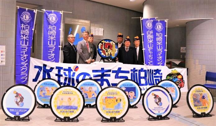 市長と教育長とライオンズクラブの方々が水球に関する絵柄の異なる9枚のマンホールの後ろに並んでいる。