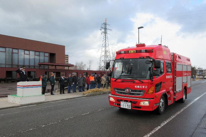 曇天の中、道路を走る赤い消防車に制服を着て敬礼のポーズをとる櫻井市長ら