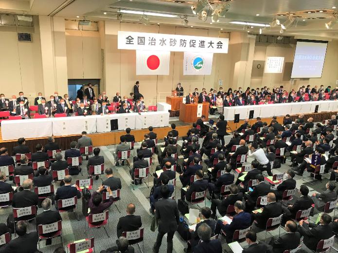 全国治水砂防促進大会と記された横看板、演壇、モニターがある体育館のような広い空間で、関係者が一堂に会している写真