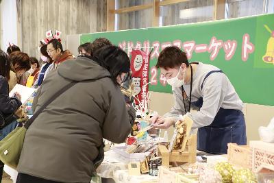 写真：お客さんに商品の説明をする男性。テーブルの上にはたくさんのハンドメイド商品が並んでいます。