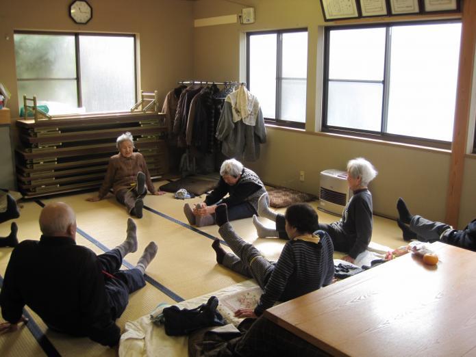 和室にてお年寄りが足を上げたり、重心を前に倒したりして体操をしている写真