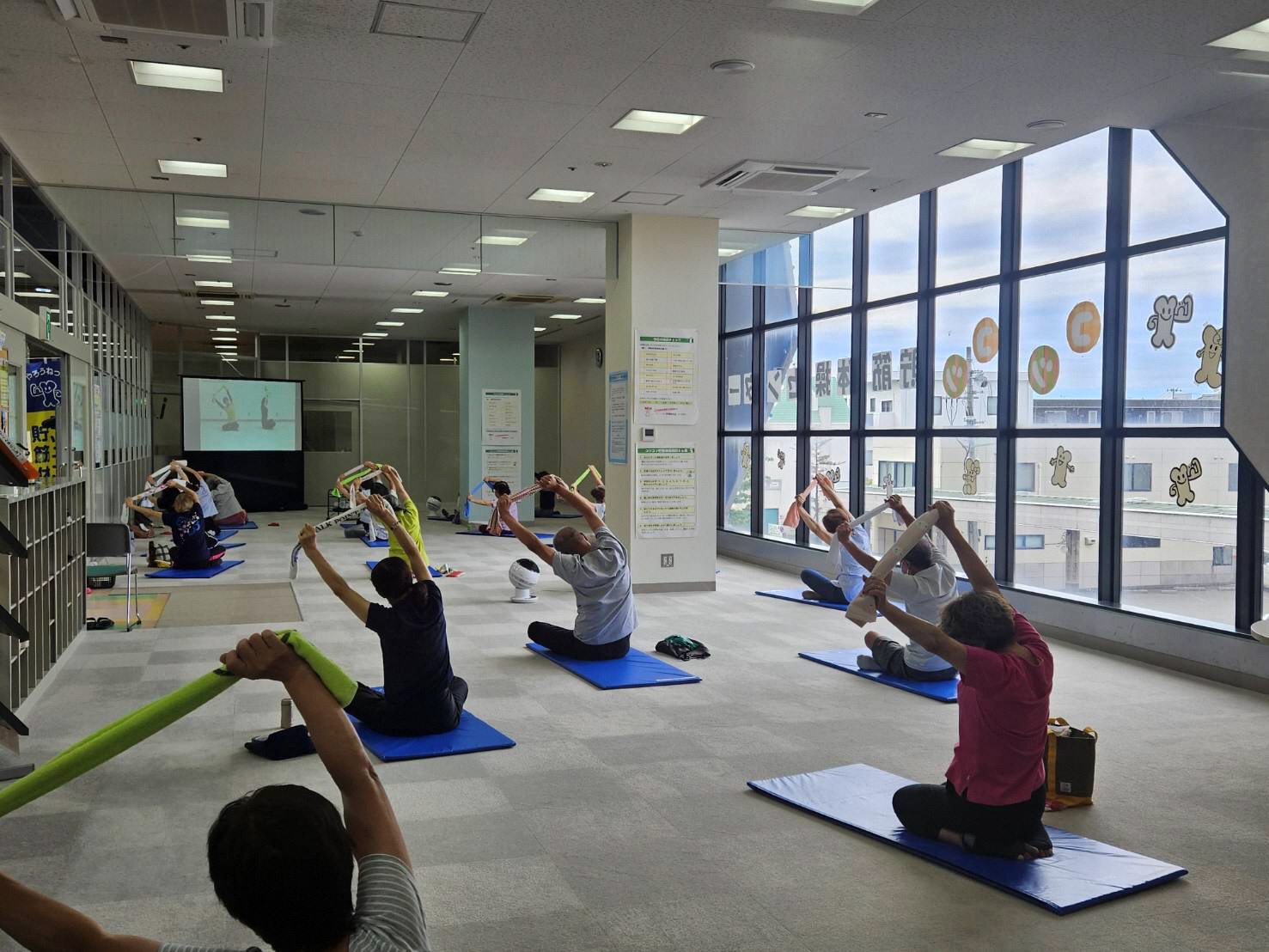 写真：体操中の利用者の皆さん。足を延ばして床に座り、タオルの両端をもって上半身を左にひねっています。