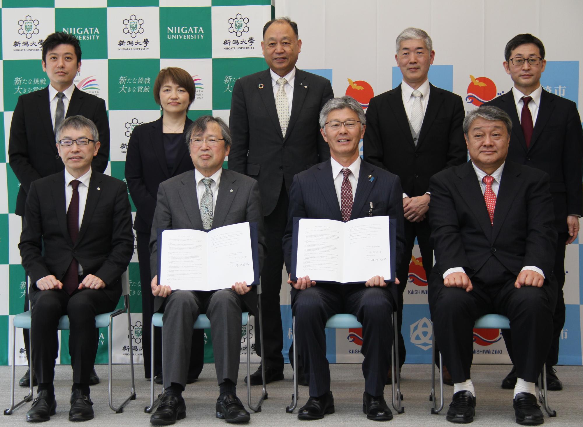 写真：署名した協定書を手に持つ市長と学長。その両脇に1人ずつ、後ろに5人が並び、記念のカメラに収まりました。