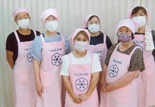 ピンク色のエプロンと三角巾をを身に着けた二中地区食生活改善推進員の女性8名中3名が前方に、後方に5名立っている写真