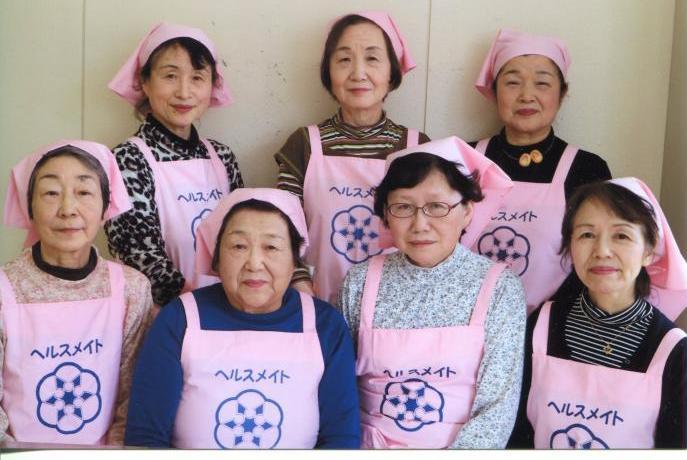 ピンク色のエプロンと三角巾をを身に着けた一中食生活改善推進員の女性7名が前方に4名、後方に3名立っている写真