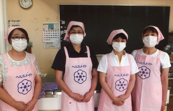 写真：ヘルスメイトと書かれたおそろいのピンク色エプロンを身に着けた4人の女性