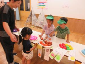 写真：小さいクラスの子がアイスのコーンを持ち、5歳児のアイス屋さんがトングでアイスをのせてあげています。