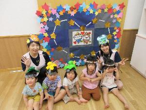 写真：2歳児と3歳児の子どもたちが織姫や彦星の帽子をかぶり完成した七夕の絵の前に並んで座っています。