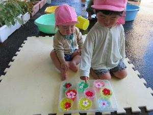 写真：1歳児の子どもたちが日陰に敷いたマットの上に座り色水に花を浮かべて見ています。
