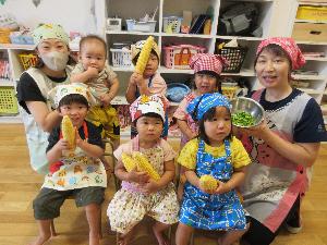 写真：エプロンを身に着けた子どもたちが皮むきしたトウモロコシを持って並んでいます。