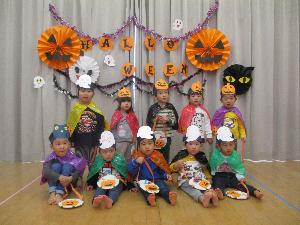 写真：ハロウィンの飾りの前でみんなで一緒に写真を撮っているぱんだ組の2歳児。子どもたちの頭には、カボチャやお化けのお面をかぶっています。