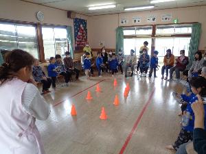 写真：三角コーンの周りに子どもと利用者が座っています。利用者の皆さんと作った新聞紙の輪を使って、輪投げ競争をしようとしている5歳児