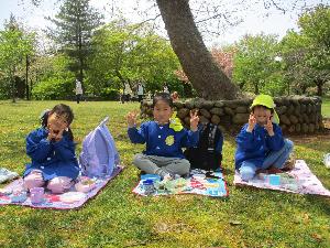 写真：レジャーシートの上に座ってお弁当を広げるらいおん組の3人の子どもたち。飛び切りの笑顔でピースサインをしてくれました。