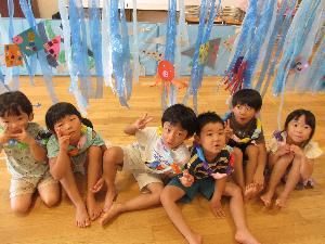 写真：水族館に見立てた部屋で子どもたちが並んで座っています。天井からたくさんのスズランテープがぶら下がり、画用紙でできた海の生き物が貼られています