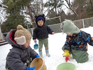 写真：未満児3人がスコップとバケツを持って雪遊びをしています