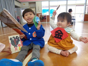 写真：金太郎の絵本を選んだ2人の子どもがいます。小さいクラスの子どもが「金」の文字がかかれた胸当てをつけ、紙でできたまさかりを持っています。