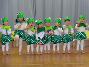 写真：かえるの帽子をかぶり、緑色のスカーフとカボチャパンツを着た2歳児・3歳児の8人が、かえるの歌を歌っています