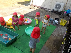 写真：建物の日陰に水の入ったたらいをいくつか置き、水遊びを楽しむりす組の子どもたち