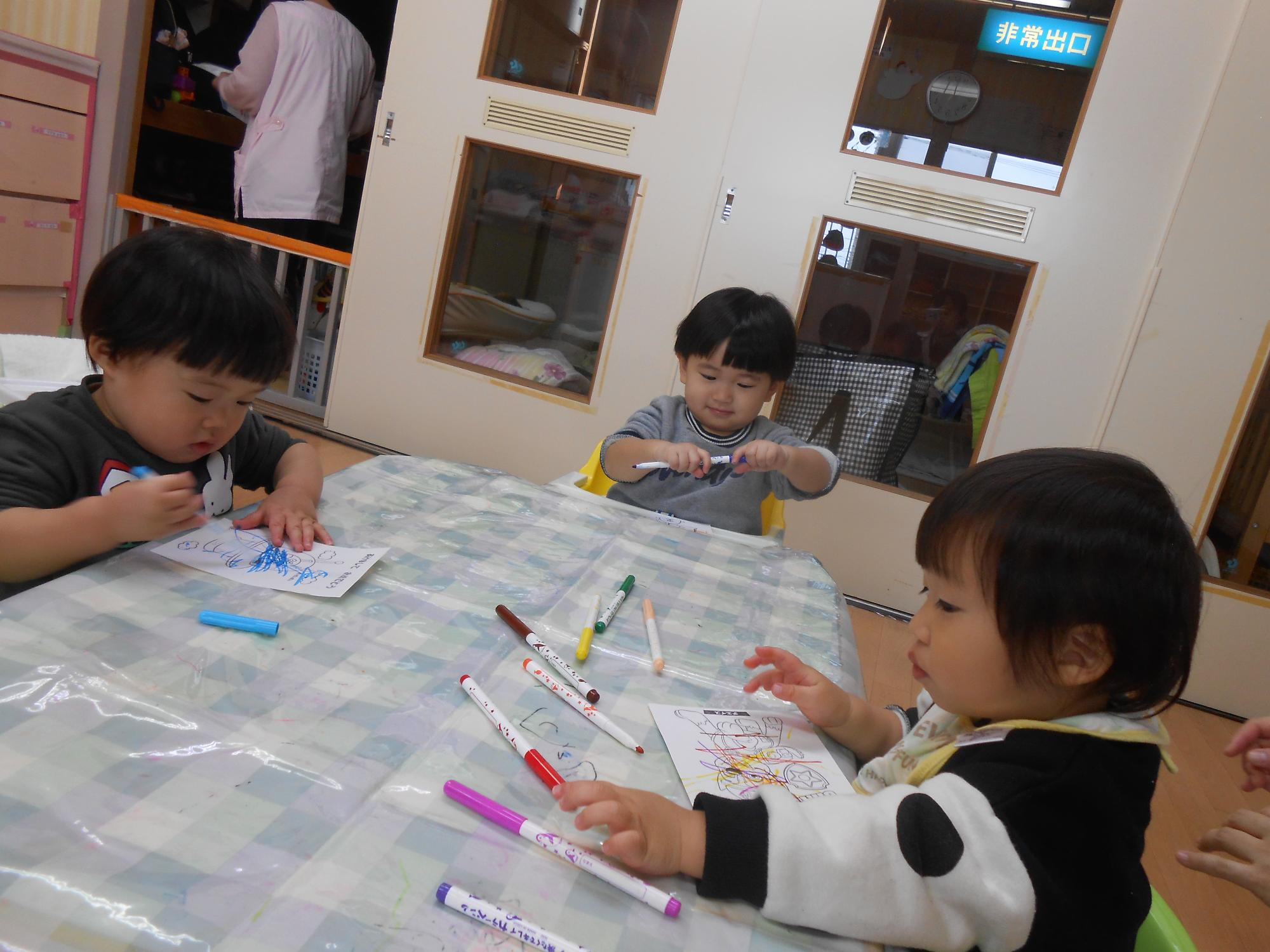 写真：0歳児の子ども達が、椅子に座ってカラーペンで年賀状に色塗りをしているところ