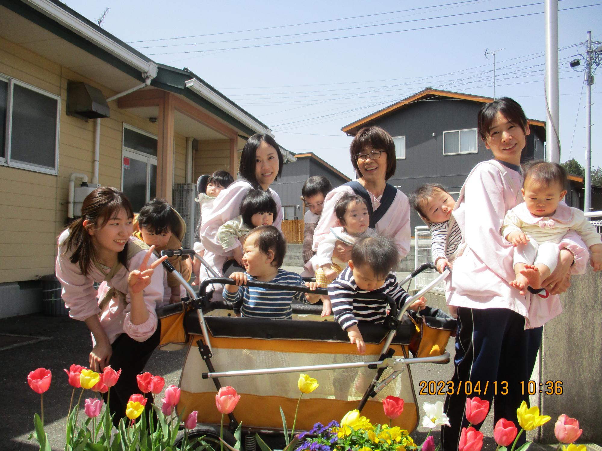 写真：チューリップの咲くプランターの後ろに並ぶ0・1歳児と保育士たち