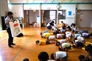 写真：お遊戯室に集まった子どもたちが、講師の合図でダンゴムシのように小さく丸まって、両手で頭を隠すポーズをしています。