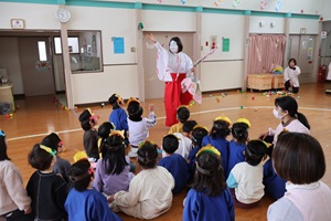 写真：鬼を退け、福の神を迎える子どもたち。座った子どもたちのに、お面をつけた福の神が花びらをふわっと撒いています