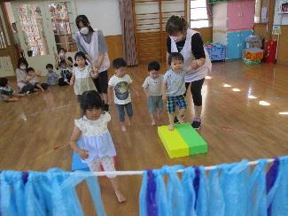 ソフト積み木の橋を渡って川に見立てたスズランテープののれんをくぐろうとする5名の1,2歳児