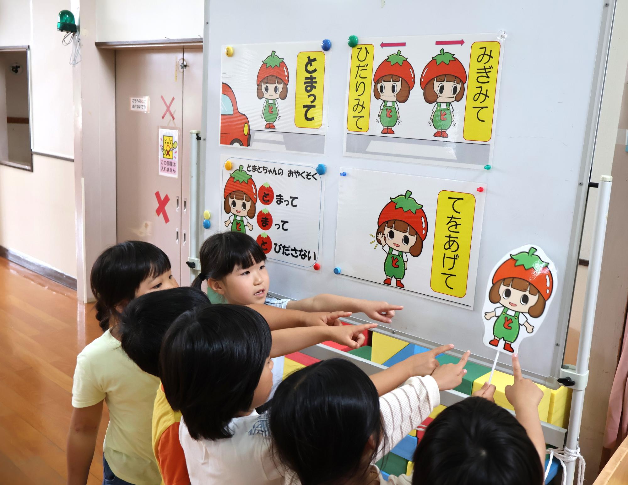 写真：トマトの被り物をした女の子のキャラクターと交通安全の約束をホワイトボードに掲示してあり、それを指をさして子どもたちが確認しています