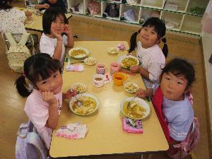 写真：年長児が作ったカレーをおいしそうに食べている4歳児と5歳児の子どもたち。カメラ目線でニッコリ笑顔を見せてくれました。