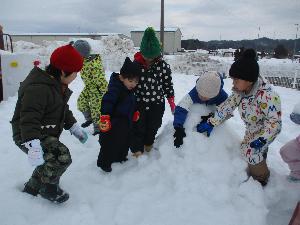 写真：雪の積もった園庭で、年長児の男の子4人と1歳児の男の子がかまくらを作っているところ