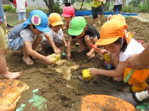 きりん組の男児2名とうさぎ組の女児3名、りす組の女児1名が砂場でプラスチックコップに入れた泥水をかき混ぜています。