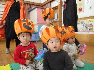 写真：画用紙で作ったかぼちゃの帽子をかぶった1歳児。視線の先には仮装した4・5歳児がいます