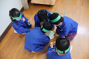 写真：5人の子どもたちが、頭を寄せ合ってピザの台紙に具材を載せているところ。