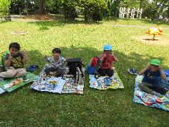 写真：芝生の上にそれそれレジャーシートを広げて座り、友達と嬉しそうにお弁当を食べている4・5歳児。