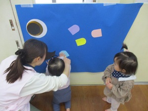 写真：1歳児の子どもが画用紙で作ったうろこを青色の大きなこいのぼりの用紙に貼っています