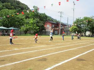 青と赤の衣装をつけた子どもたちが、グラウンドで1列に並び、キラキラテープがついた棒を持って踊っている様子