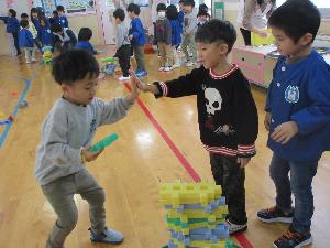 写真：ブロック積みゲームをしている子どもたち。3人の子どもたちの前に積み上げられたブロックがあり、次に積み上げる子にタッチをしています。