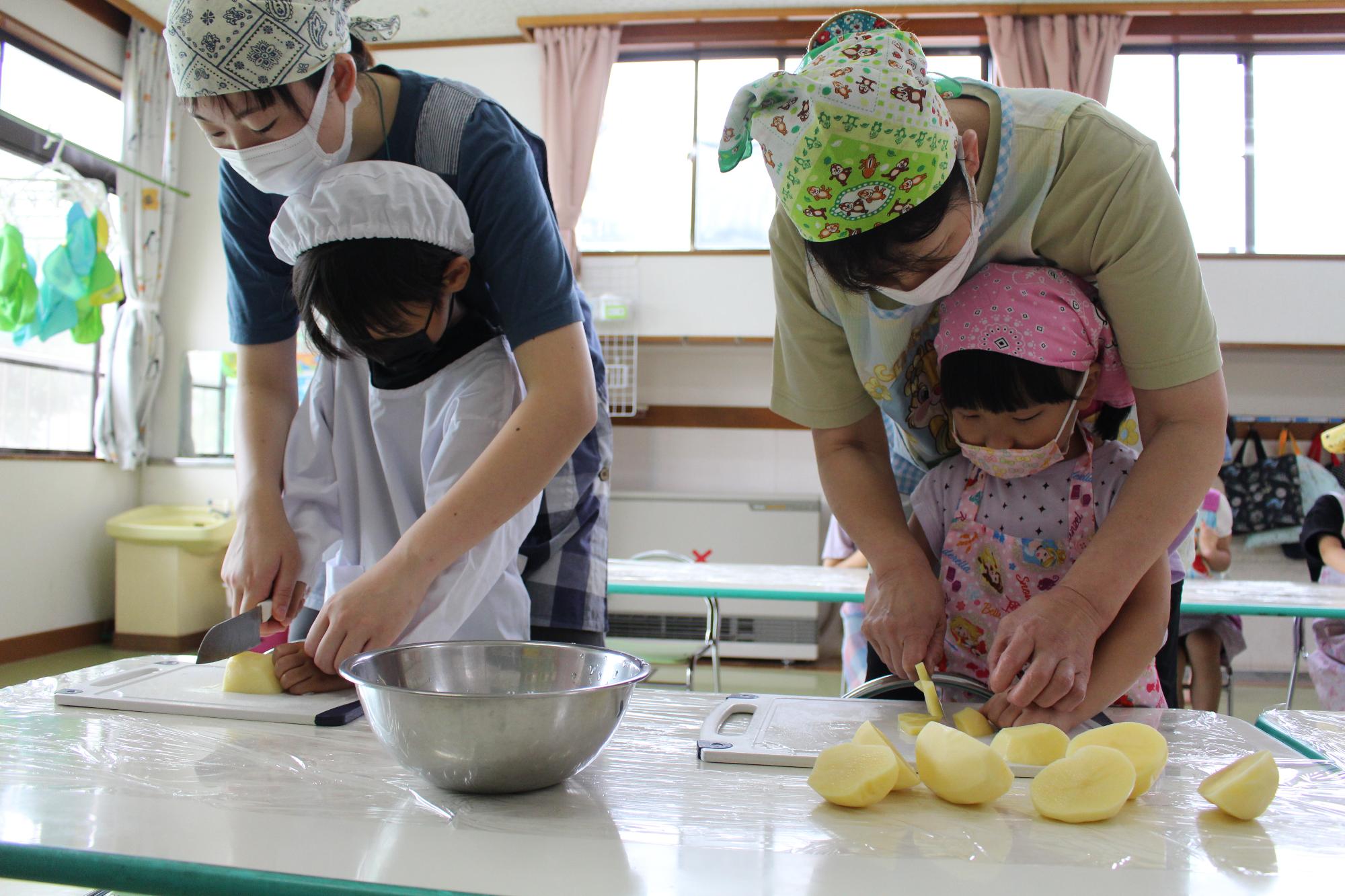 写真：年長児と年中児がエプロンを着て包丁でジャガイモを切っている様子。保育士が後ろから手を添えています。
