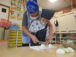 写真：包丁を持った年長児の男の子が職員に手を添えられながらタマネギを切っている