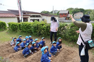 写真：畑の畝の間に子どもたちが並び、保育者が苗の植え方を教えているところ