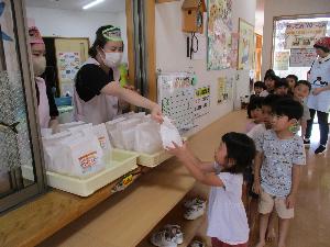 写真：保育園の廊下に一列に並んだらいおん組の子どもたちが、事務室の窓から紙袋に入ったサバサンドを受け取っているところ