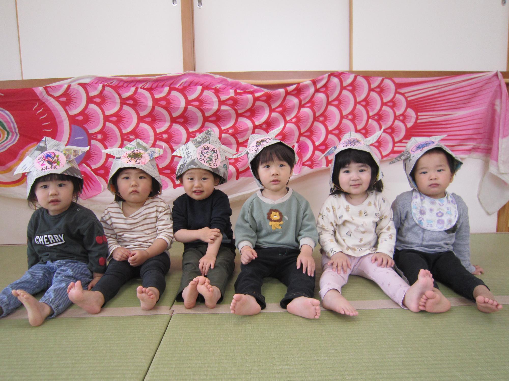 写真：壁に飾った大きなこいのぼりの前に並んで座る6人の子ども