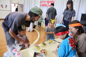 写真：机の上にたくさん並んだ食べ物やおもちゃを親子で買い物をしているところ