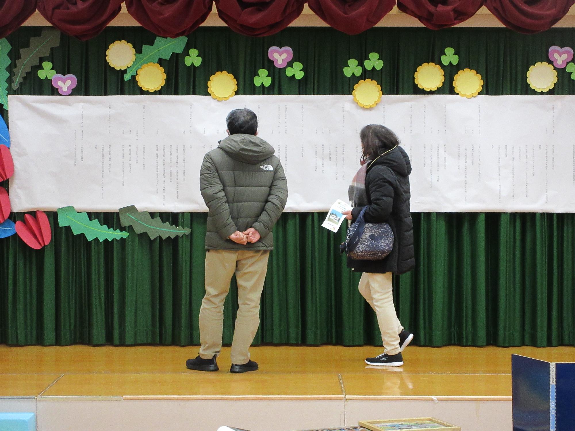 写真：ステージの横幅いっぱいに展示された高柳保育園の沿革。年配のご夫婦が見ています。